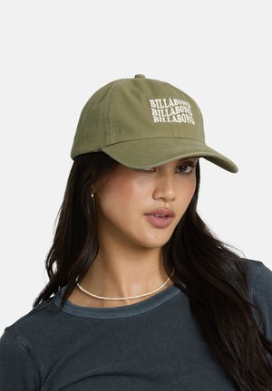 Giovane donna con capelli lunghi e scuri che indossa un cappellino verde Billabong, piccoli orecchini a cerchio, una collana di perline e una maglia grigio scuro.