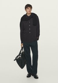 Zwarte button-up shirt, donkere broek en zwarte schoenen. Een zwarte trui over de schouders draped. Het model houdt een zwarte tas vast.