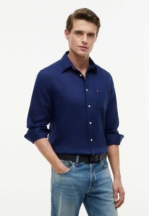 Mann trägt ein marineblaues Hemd mit langen Ärmeln und hochgekrempelten Ärmeln sowie hellblaue Jeans, steht mit einer Hand in der Tasche.