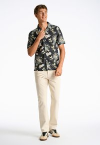 Camicia nera a maniche corte con motivo floreale, realizzata in tessuto leggero, abbinata a pantaloni color crema e sneakers nere e beige.