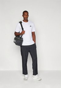 Camisola branca de manga curta com logótipo preto, combinada com calças escuras e sapatilhas desportivas brancas. Saco de ombro preto com texto branco.