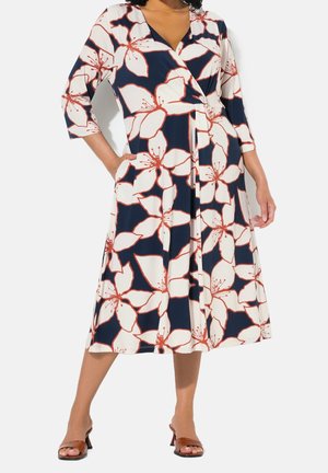 Kobieta w granatowej sukience kopertowej z dużym białym i pomarańczowym kwiatowym wzorem, z rękawami o długości 3/4 i brązowych sandałach na wysokim obcasie.