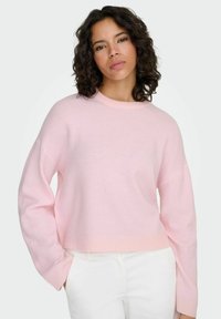 Light pink cropped sweater with long sleeves and a round neckline, made from a soft, textured fabric. Worn over white pants.