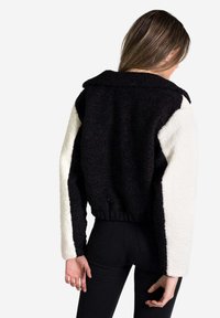 Chaqueta recortada en blanco y negro con una textura esponjosa, que presenta un cuello y un dobladillo elástico. Cuerpo negro y mangas blancas.
