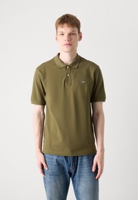 Olivengrønt polo-shirt med to knapper, korte ærmer og subtilt logo; parret med blå jeans for et afslappet look.