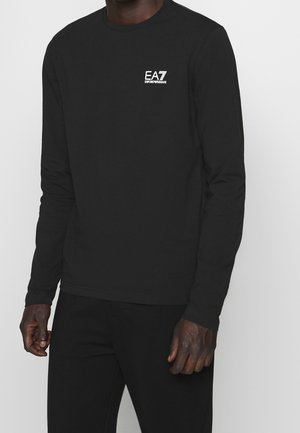 Mies pukeutuneena mustaan EA7 Emporio Armani -merkkiseen pitkähihnaiseen paitaan, jossa rintalogo, sekä mustiin housuihin, seisoo yksinkertaisen vaalean taustan edessä.