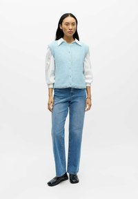 Lys blå strikket vest med v-hals og knappelukking, båret over en hvit skjorte med puffede ermer, kombinert med blå jeans med rette ben og svarte sko.