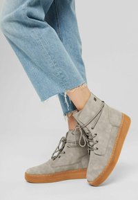 Bottes chevilles en daim gris avec une semelle en caoutchouc marron, un design à lacets et des coutures contrastantes, associées à un jean bleu clair déchiré.