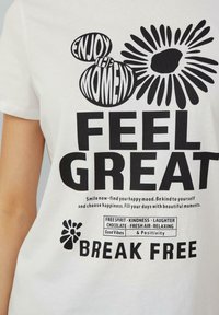 Witte katoenen T-shirt met bold zwarte tekst: "VOEL JE GOED" en "BREK UIT," met decoratieve grafische elementen zoals bloemen en bellen.