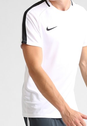 Homme portant un maillot de sport Nike blanc avec des bandes noires sur les épaules, debout devant un fond clair uni.