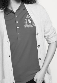 Polo rouge avec col, présentant un logo brodé blanc, superposé sous un cardigan en tricot gris clair à boutons.