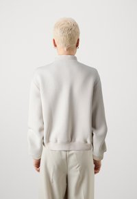 Helles graues Sweatshirt mit Rundhalsausschnitt, weichem Material und gerippten Bündchen, getragen mit beigen Hosen, von hinten betrachtet.