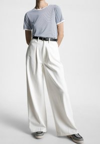Camisa de manga corta a rayas azul y blanca, pantalones anchos blancos de talle alto, cinturón negro y zapatillas grises con patrones texturizados.