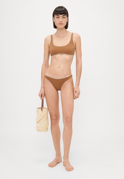 Rudas bikinis, pagamintas iš lygios medžiagos, su baltais zigzagais aplink kraštus. Modelis laiko apvalią pintą šiaudų rankinę.