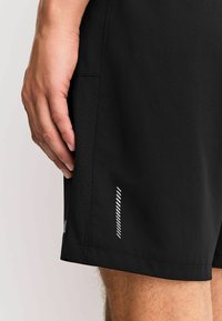 Schwarze Sportshorts mit glatter Textur, ausgestattet mit einer Seitenpaneel mit Perforationen und einem weißen gestreiften Akzent nahe dem Saum.