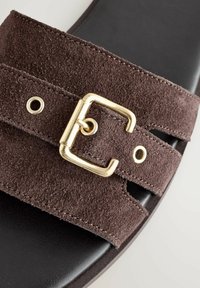 Sandale en daim marron avec des grosses sangles, dotée d'une boucle en or et d'un design découpé. Semelle noire à texture lisse, mettant en valeur les sangles.