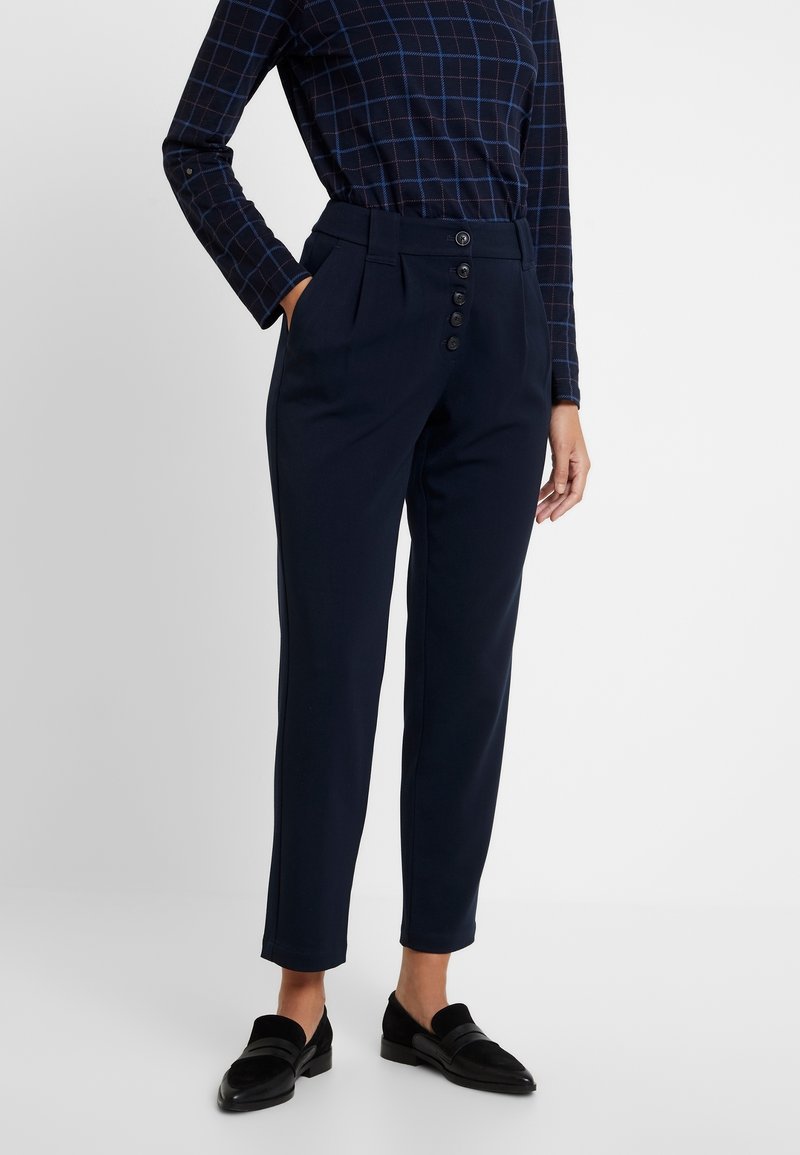 Granatowe spodnie o wysokim stanie, z fałdami z przodu i zapięciem na pięć guzików. Połączone z ciemną koszulą w kratę z długim rękawem i czarnymi loafersami.