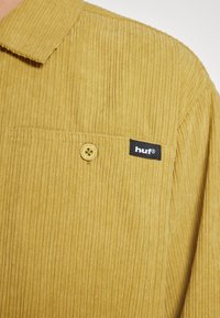 Camicia in corda gialla senape con bottoni sul davanti, caratterizzata da una tasca e un'etichetta nera con la scritta "huf". È evidente la texture a coste.