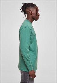 Camiseta de algodón verde de manga larga con cuello redondo, textura suave, corte relajado y dobladillo recto. Llevada con jeans grises.
