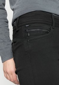 Close-up de uma pessoa a usar jeans pretos com um bolso frontal com fecho e uma camisa de mangas longas cinza, com a mão descansando ao lado da anca.