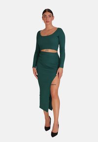 Grünes geripptes Zweiteiler-Set mit einem cropped-langärmligen Oberteil und einem Rock mit hohem Schlitz, das einen figurbetonten Schnitt und strukturierte Stoffe aufweist. Schwarze High Heels vervollständigen den Look.