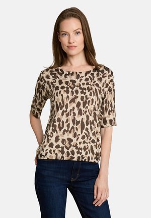 Femme aux cheveux bruns raides portant un haut beige et marron foncé à imprimé léopard à manches courtes et un jean bleu foncé, debout avec une main sur la hanche.