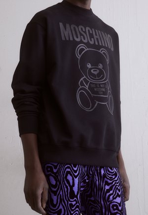 Osoba ubrana w czarną bluzę z napisem „MOSCHINO” oraz grafiką misia z napisem „This is not a Moschino toy”, w połączeniu z fioletowymi spodniami w wzory.