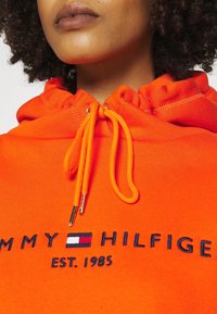 Fel oranje hoodie met een capuchon met trekkoord, met het geborduurde logo "TOMMY HILFIGER" en "EST. 1985" in donkerblauw, en zilverkleurige hardware accenten.