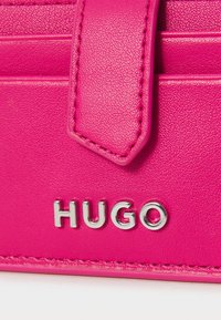 Izdelek iz roza usnja z teksturo, ki vključuje srebrni logotip "HUGO" in usklajene šive.