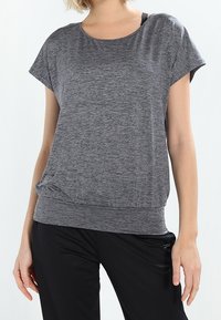 Mujer vestida con una camiseta de mangas cortas gris y pantalones negros, de pie con los brazos relajados a los lados contra un fondo claro y liso.