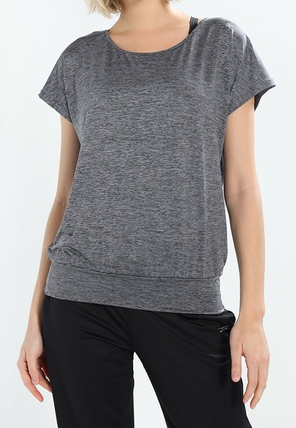 Mujer vestida con una camiseta de mangas cortas gris y pantalones negros, de pie con los brazos relajados a los lados contra un fondo claro y liso.