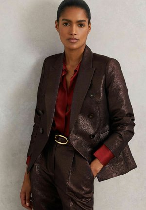 Dobbeltradet blazer i mørkebrunt med en blank tekstur, kombineret med en burgundy skjorte. Fremhævet med sorte knapper og en guld bæltespænde.