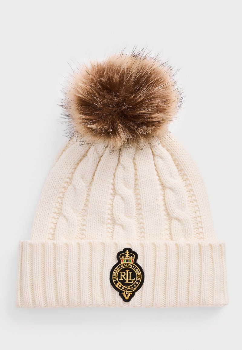 Kremasta pletiva kapa s uzorkom u obliku kabla, veliki smeđi krzneni pom-pom na vrhu i crna i zlatna oznaka Ralph Lauren na preklopu.