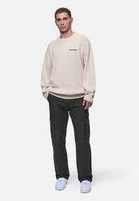 Beige gebreide trui met ronde hals en logo op de borst, gecombineerd met donkere cargobroek met zakken en witte sneakers. Schone achtergrond.
