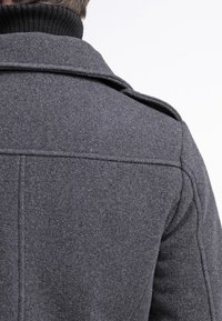 Szary wełniany płaszcz o wyraźnej fakturze, z wysokim kołnierzem i pagonami na ramionach. Tył dopasowany, z czystą, nowoczesną sylwetką.