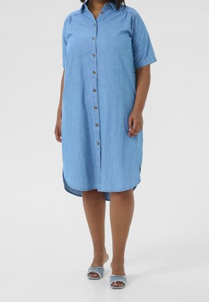 Kvinde iført en lyseblå denim kjole med knapper og korte ærmer samt slidte sandaler med åben tå, stående på en enkel lys baggrund.