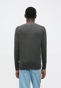 Μαύρος άνδρας με κοντά σγουρά μαλλιά, φορώντας ένα σκούρο γκρι μακρυμάνικο πουλόβερ και ανοιχτό γαλάζιο τζιν, στέκεται γυρισμένος με την πλάτη προς τον φακό, μπροστά σε λευκό φόντο.