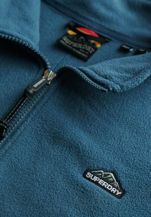 Niebieska kurtka polarowa z przednim zamkiem błyskawicznym, haftowanym logo Superdry na piersi oraz oznakowaną metką wewnątrz kołnierza.