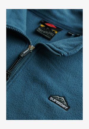Blauwe fleecejas met een ritssluiting aan de voorkant, geborduurd Superdry-logo op de borst en een merklabel aan de binnenkant van de kraag.