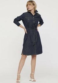 Robe chemise en denim à manches longues, col rabattu, poches avant, fermeture à boutons, taille ceinture et fentes latérales. Couleur bleu foncé.
