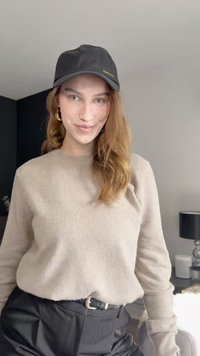 Bézs színű, texturált hosszú ujjú pulóver, magas derekú fekete nadrággal és arany logót tartalmazó fekete sapkával viselve.