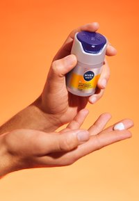 Crema idratante Nivea Men in una bottiglia grigia e blu navy, tenuta in una mano, con una piccola quantità di crema bianca sul palmo. Sfondo arancione.