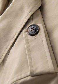 Beżowy materiał o gładkiej fakturze, z czarnym guzikiem na zapięciu klapy, widocznymi detalami przeszyć oraz wyraźnymi klapami.