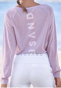 Długi rękaw, jasnofioletowa koszula o luźnym kroju, z pionowym białym napisem "SAND" na plecach. Do tego białe, dopasowane spodnie.