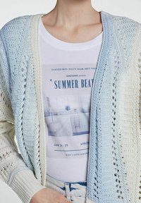 Crochet-neulecardigan vaaleansinisen ja valkoisen liukuvärin sävyissä, avoin etuosa, jossa on monimutkaisia kuvioita. Alla valkoinen T-paita, jossa on graafinen printti.