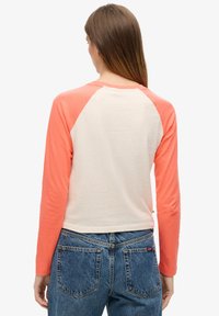 Woman wearing long-sleeve raglan shirt with cream body and coral sleeves, paired with blue denim jeans, facing away from the camera.