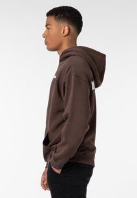 Brauner Kapuzenpullover mit einer Fronttasche, einfarbig und mit gerippten Bündchen. Verfügt über eine verstellbare Kapuze und weißen Text auf der Brust.