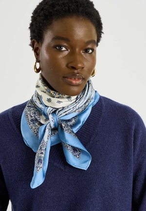 Femme aux cheveux courts et bouclés portant des boucles d'oreilles créoles en or, un pull bleu, et un foulard en soie bleu et blanc à motifs noué autour du cou.