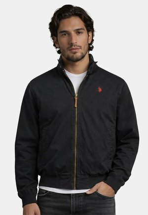 Mann trägt schwarze Jacke mit Reißverschluss über weißem Hemd mit rotem besticktem Logo, steht vor einfarbig hellem Hintergrund.