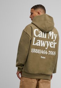 Olivengrønn hettegenser laget av myk stoff, med stor hvit tekst på baksiden: "Ring min advokat" og et telefonnummer. Hette-design, løs passform.